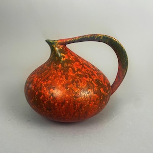 Puede incluir: Jarra de cerámica con cuerpo bulboso y asa curva. La jarra es principalmente naranja con una textura moteada, que pasa a marrón y verde cerca del asa. Una pieza de cerámica única.