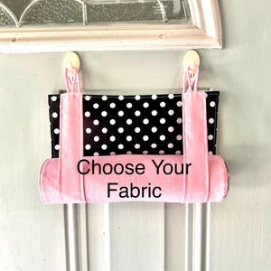 May include: A fabric hanging organizer featuring a black rectangular panel with white polka dots and a solid light pink cylindrical pocket at the bottom. The unit has two light pink fabric straps and displays the text Choose Your Fabric.