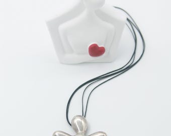 Puede incluir: Una escultura de cerámica blanca de dos figuras abrazándose, con un detalle de corazón rojo. Un colgante plateado con forma de flor cuelga de cordones negros, sugiriendo un tema de amor y relaciones. El colgante es el punto focal.