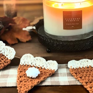 May include: A lit, three-wick candle in a glass jar with a label reading "Cashmere Pumpkin" sits on a black metal tray. Crochet pumpkin pie slice garland decorates the table, creating a cozy autumn theme.
