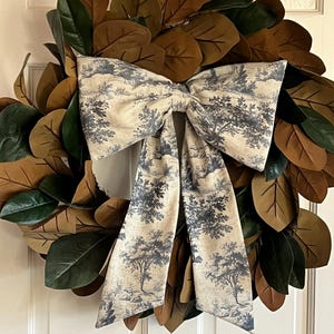 May include: A decorative wreath featuring large, brown and green leaves. A large bow made of fabric with a blue and white landscape pattern is centered on the wreath. The wreath is hanging on a white door.