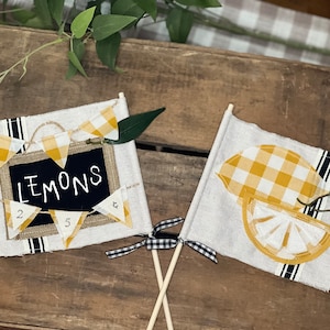 May include: Two fabric flags with wooden sticks. The first flag is white with a black and white striped border and a yellow and white gingham lemon design. The second flag is white with a black and white striped border and a chalkboard sign that says "Lemons" with yellow and white gingham bunting.