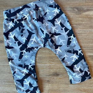 Puede incluir: Pantalones de bebé grises y negros con un diseño de camuflaje de aviones. Los pantalones tienen una cintura elástica con cordón y piernas con puños.