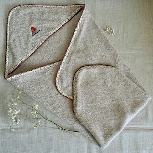 Peut inclure: Une serviette à capuche beige en tissu éponge avec une bordure à motif floral rouge foncé et blanc. La capuche présente un petit motif brodé en forme de cœur rouge. La serviette est pliée pour montrer la capuche et le tissu texturé.