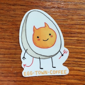 May include: A white sticker featuring a cartoon egg character with a devilish orange yolk, horns, and a red pitchfork. The egg has a smiling face and a red tail. The text "EGG TOWN COFFEE" is at the bottom.