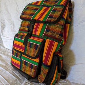 May include: A black backpack with a colourful patterned fabric design. The fabric is a mix of yellow, red, green, and black stripes and squares. The backpack has a large main compartment, a smaller front pocket, and two side pockets.