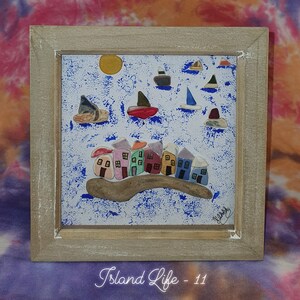May include: A framed artwork showing a coastal scene with colourful houses, boats, and a sun against a blue and white background. The frame is light-coloured wood. The text "Island Life - 11" is at the bottom of the artwork.