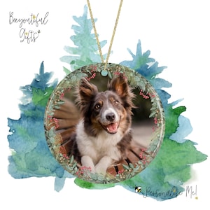May include: A round, white, ceramic ornament with a festive wreath design featuring green leaves, red berries, and a brown wooden background. A brown and white dog with a happy expression is pictured in the center of the ornament. The ornament is hanging from a gold chain.