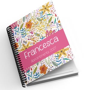 May include: A spiral-bound appointment book featuring a white cover with a colorful floral pattern of yellow, orange, pink, and blue botanical elements. A solid pink horizontal band displays the white text Francesca and Appointments 2027.