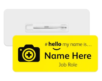 May include: A yellow name tag with a black camera icon and the text "#hello my name is... Name Here Job Role." The tag has a white backing with a pin for attachment.