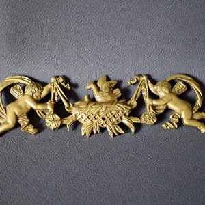 Op de afbeelding: Gouden metalen ornament met twee engelen die linten vasthouden en een centrale vogel in een nest. Het ornament is gedetailleerd met ingewikkelde gravures en een delicaat ontwerp.