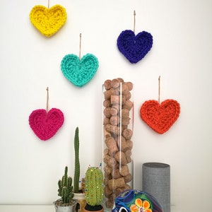 May include: Five crocheted hearts in different colors hanging on a white wall, with a glass vase filled with cork stoppers, two potted cacti, a painted sugar skull, and a gray smart speaker on a white table.