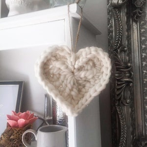 May include: A white crocheted heart hanging from a white shelf. The heart is made of thick yarn and has a textured surface. The shelf is decorated with a white skull and a clear glass jar.