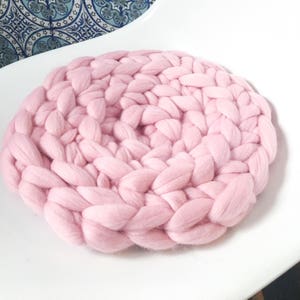 May include: A large, round, pink knitted cushion. The cushion is made of thick, chunky yarn and has a soft, fluffy texture.