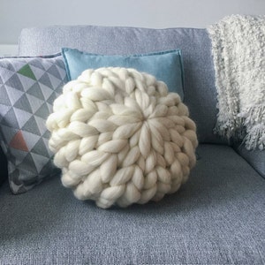 May include: A large, white, chunky knit throw pillow on a gray couch. The pillow is made of thick yarn and has a round shape.