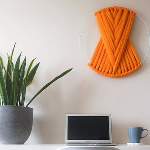 May include: A white wall with a large orange yarn wall hanging, a potted snake plant, a laptop computer, and a gray mug on a white surface.