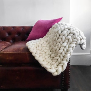 May include: A white chunky knit throw blanket draped over a brown leather sofa with a pink pillow.