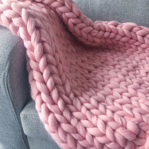 May include: A soft, pink chunky knit throw blanket draped over a gray couch. The blanket is made of thick yarn and has a loose, textured weave.