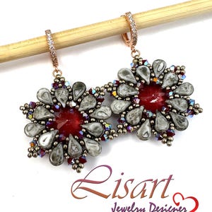 Può includere: Un paio di orecchini di perline con un cristallo rosso centrale circondato da perline grigie a forma di goccia e piccoli accenti rosso scuro. Gli orecchini pendono da cerchi color oro rosa con piccole pietre chiare. Testo: Lisart Jewelry Designer.
