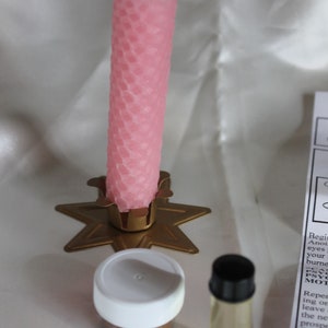 May include: A pink honeycomb-patterned candle stands in a gold star-shaped holder. Beside it are a small jar with a white lid and a clear bottle with a black cap. The items are on a white surface.