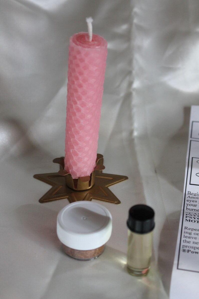 May include: A pink honeycomb-patterned candle stands in a gold star-shaped holder. Beside it are a small jar with a white lid and a clear bottle with a black cap. The items are on a white surface.