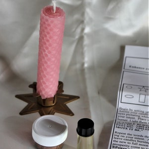 May include: A pink honeycomb-patterned candle stands in a gold star-shaped holder. Beside it are a small jar with a white lid, a small glass bottle with a black cap, and a printed instruction sheet. The items are arranged on a white surface.