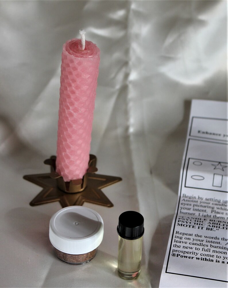May include: A pink honeycomb-patterned candle stands in a gold star-shaped holder. Beside it are a small jar with a white lid, a small glass bottle with a black cap, and a printed instruction sheet. The items are arranged on a white surface.