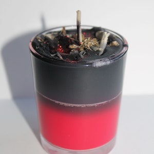 May include: A layered candle in a clear glass. The bottom half is a vibrant red, transitioning to black at the top. The top is covered with black material, dried herbs, and a partially burned wick.