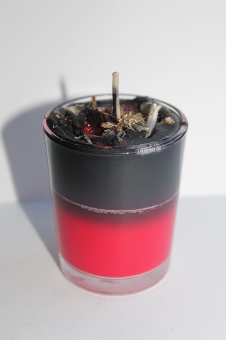 May include: A layered candle in a clear glass. The bottom half is a vibrant red, transitioning to black at the top. The top is covered with black material, dried herbs, and a partially burned wick.