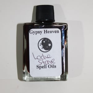 May include: A small, clear glass bottle filled with a dark, shimmering liquid. The bottle has a black cap and a white label with the text "Gypsy Heaven" and "Lodie Stone Spell Oils". A crescent moon and stars graphic is also on the label.
