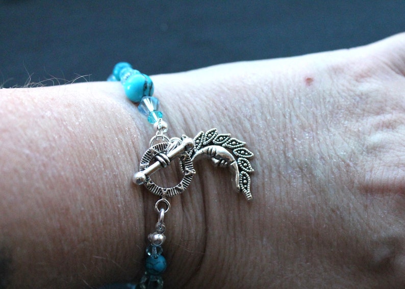 May include: A turquoise and silver bracelet with a decorative clasp featuring a crescent moon and leaves. The bracelet is made with turquoise beads and silver beads.