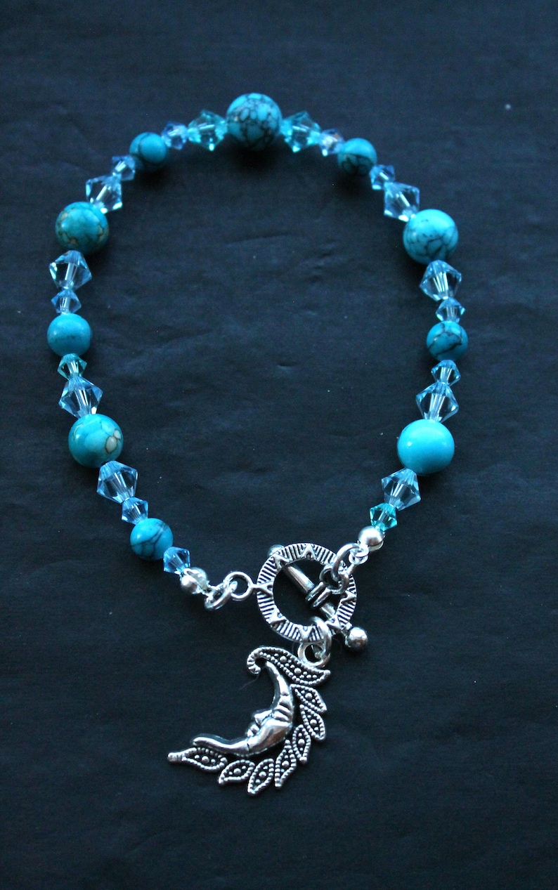 May include: A turquoise and silver bracelet with a crescent moon charm. The bracelet is made with turquoise beads and clear crystal beads.