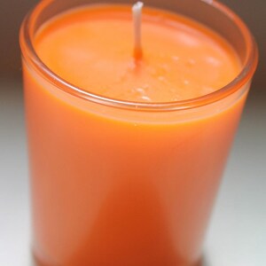 May include: An orange candle in a clear glass container. The candle is a solid color with a white wick centered in the wax. The glass is cylindrical and the candle is lit.