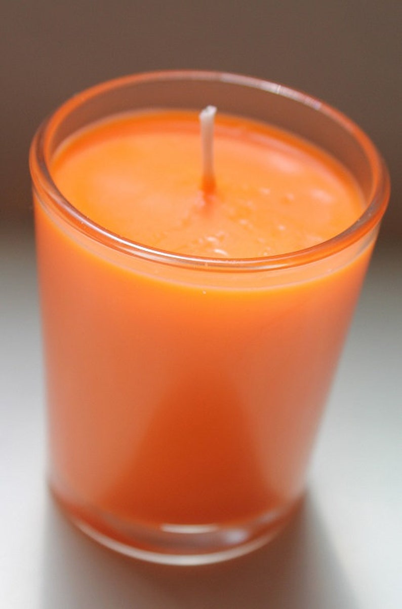 May include: An orange candle in a clear glass container. The candle is a solid color with a white wick centered in the wax. The glass is cylindrical and the candle is lit.