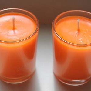 May include: Two orange-colored candles in clear glass holders. Each candle has a white wick centered in the wax. The candles are cylindrical and appear to be unscented. The glass holders have a simple, clean design.