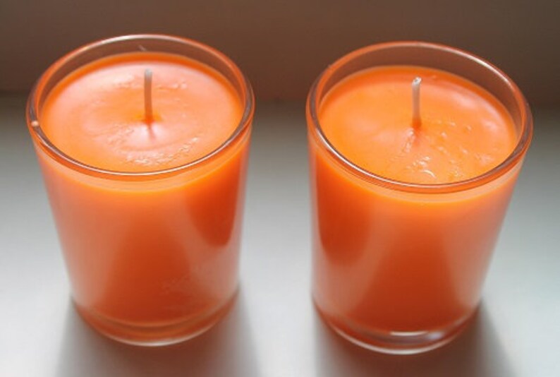 May include: Two orange-colored candles in clear glass holders. Each candle has a white wick centered in the wax. The candles are cylindrical and appear to be unscented. The glass holders have a simple, clean design.