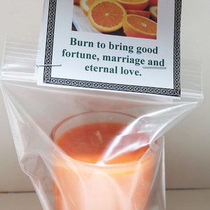 May include: Orange-scented candle in a clear glass, packaged in a plastic bag. The candle is a vibrant orange color. A tag above the candle reads "ORANGE" with an image of orange slices and the text "Burn to bring good fortune, marriage and eternal love."