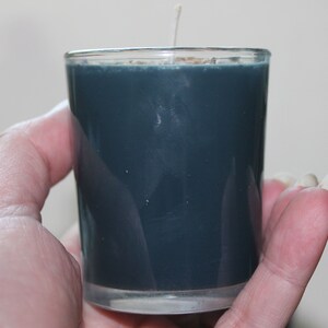 May include: A teal-colored votive candle in a clear glass holder. The candle has a white wick and is held in a hand. The candle is a solid color and appears to be unscented. The glass holder is cylindrical and has a smooth surface.