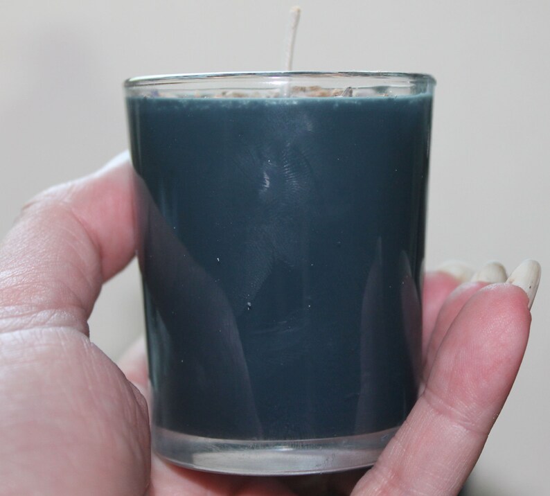 May include: A teal-colored votive candle in a clear glass holder. The candle has a white wick and is held in a hand. The candle is a solid color and appears to be unscented. The glass holder is cylindrical and has a smooth surface.