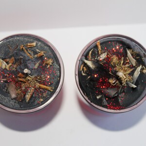 May include: Two round, black candles with silver rims. The candles are decorated with dried herbs, small pieces of wood, and red glitter. The candles are on a white surface.