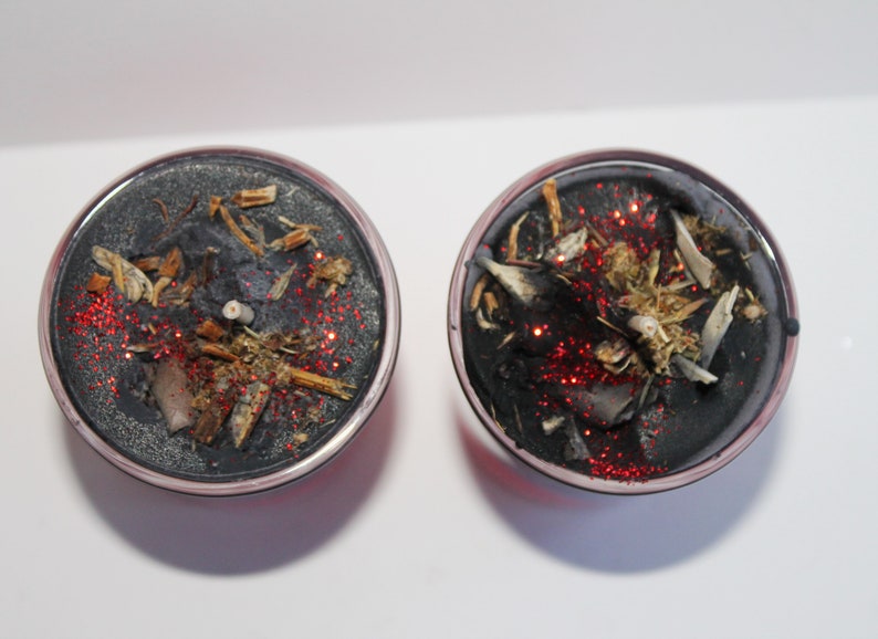 May include: Two round, black candles with silver rims. The candles are decorated with dried herbs, small pieces of wood, and red glitter. The candles are on a white surface.