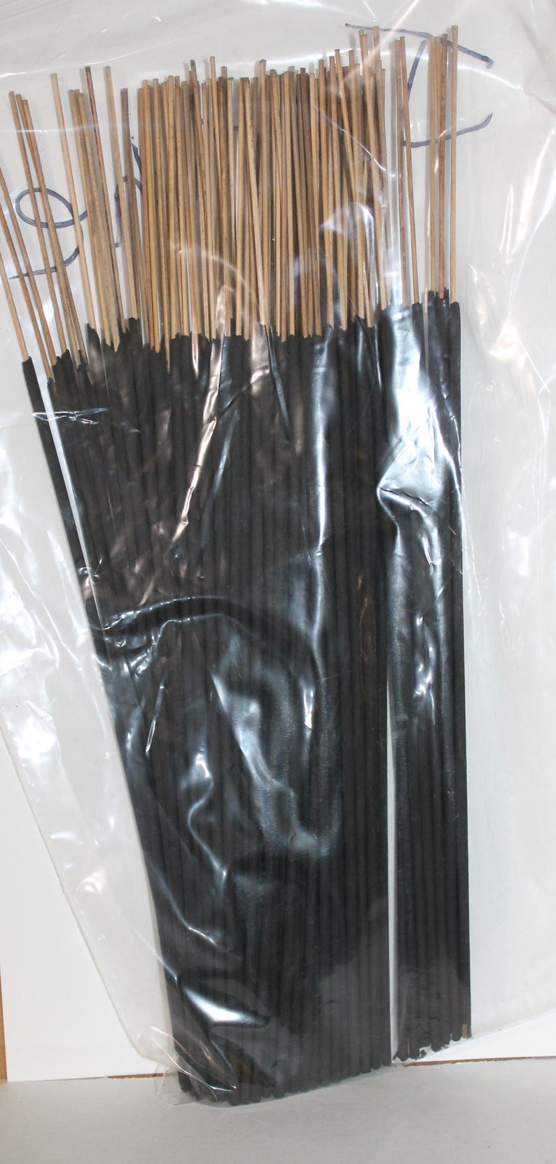 May include: A clear plastic bag filled with a bundle of black incense sticks. The sticks are tied together with a thin brown string.