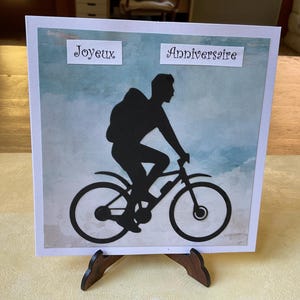 Peut inclure: Une carte de vœux carrée présentant la silhouette noire d'une personne à vélo avec un sac à dos sur un fond aquarelle bleu clair et blanc. Le texte Joyeux Anniversaire est imprimé sur deux étiquettes rectangulaires blanches séparées.