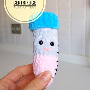 May include: A handmade amigurumi centrifuge tube in blue, gray, and pink. The plush toy has black eyes and a small pink nose. The text "AMIGURUMI CENTRIFUGE TUBE PATTERN" is in a yellow circle.