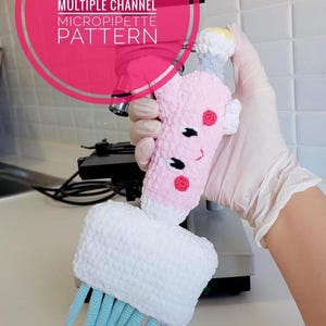 Könnte beinhalten: Eine gehäkelte Amigurumi-Mikropipette in Rosa und Weiß mit einem lächelnden Gesicht und roten Akzenten. Die Pipette hat eine weiße Basis mit blauen Häkelsträngen. Das Bild enthält den Text "AMIGURUMI MULTIPLE CHANNEL MICROPIPETTE PATTERN" und "US ENGLISH PDF".