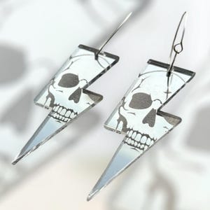 May include: A pair of silver lightning bolt shaped earrings with a skull design. The earrings are made of acrylic and have a mirrored finish.
