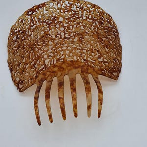 May include: A vintage hair comb with a decorative, openwork design. The comb features a semi-circular top with intricate cut-out patterns and a row of five teeth. The color is a warm, mottled brown, suggesting a tortoiseshell material. The comb is set against a white background.