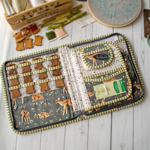 May include: A fabric organizer case featuring a fawn and floral print in shades of green, brown, and white. The interior includes elastic loops for wooden thread spools, a zippered pocket, and a sleeve containing a pack of Clover gold eye needles.