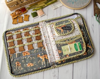 May include: A fabric organizer case with a zipper closure, featuring a dark green background with a fawn and floral print. The interior includes elastic loops holding wooden thread spools, a zippered pocket, and a sleeve containing a Clover needle pack.
