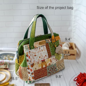 May include: A quilted project bag featuring a patchwork design of green, orange, and white autumn-themed fabrics with acorn motifs. The bag has two green handles, a zipper closure, and a hanging acorn charm. Dimensions are 9.8 inches by 9.4 inches by 5.5 inches.